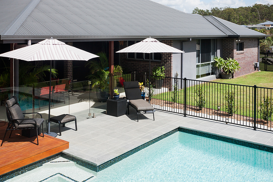 modern hotel with poolside and relaxing area with chairs on the wooden and tiled floor beside glass and metal fence there are walls made in brick which showing the roof the water brightens with sunlight